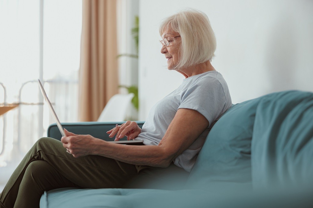 Happy senior woman sitting on modern sofa and browsing laptop computer on blurred background of her home